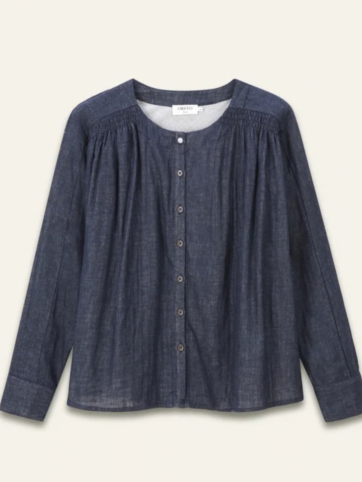 A dark blue chambray button-up blouse with a smocked neckline, long sleeves, and a relaxed fit, shown against a light cream background.