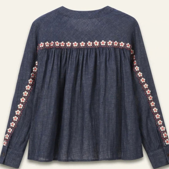 A dark blue denim long-sleeve blouse with a gathered waist and embroidered floral details along the neckline and down the sleeves, shown against a light cream background.