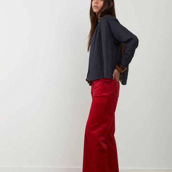 A woman stands in profile wearing a dark navy long-sleeve top and bright red wide-leg pants against a plain white wall and light wood floor.