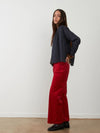 A woman stands in profile wearing a dark navy long-sleeve top and bright red wide-leg pants against a plain white wall and light wood floor.