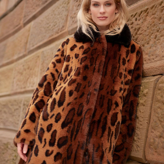 A woman wearing a brown and black leopard-print faux fur coat with a black collar, leaning against a stone wall.