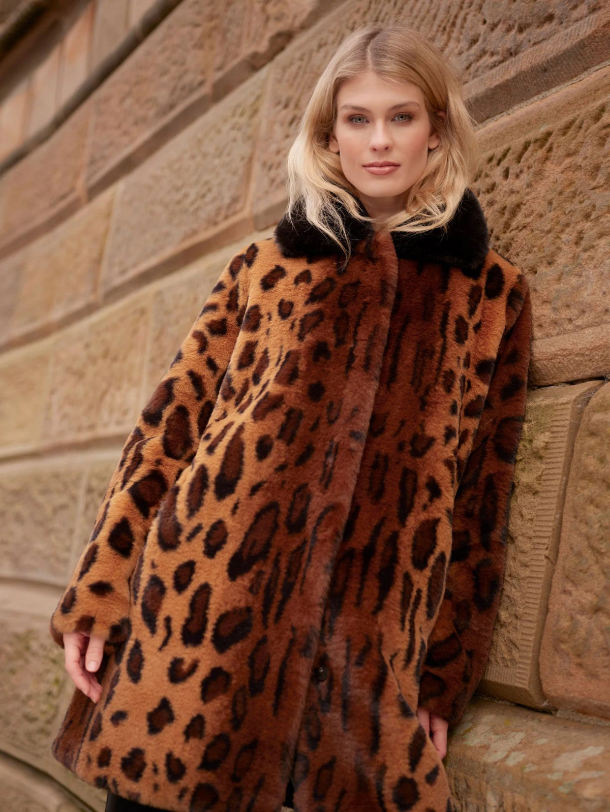 A woman wearing a brown and black leopard-print faux fur coat with a black collar, leaning against a stone wall.
