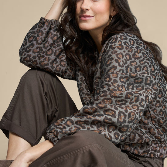 A woman wearing a leopard-print long-sleeve blouse and dark brown wide-leg pants, posing against a solid beige background.