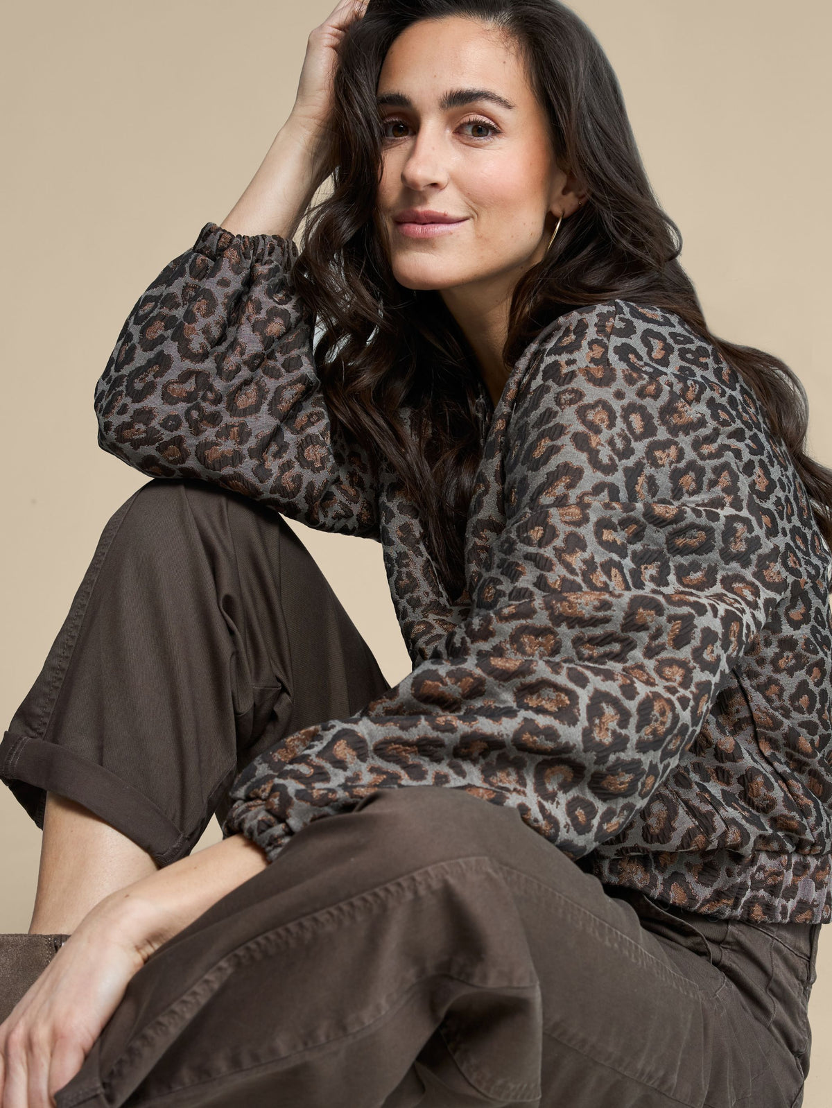 A woman wearing a leopard-print long-sleeve blouse and dark brown wide-leg pants, posing against a solid beige background.