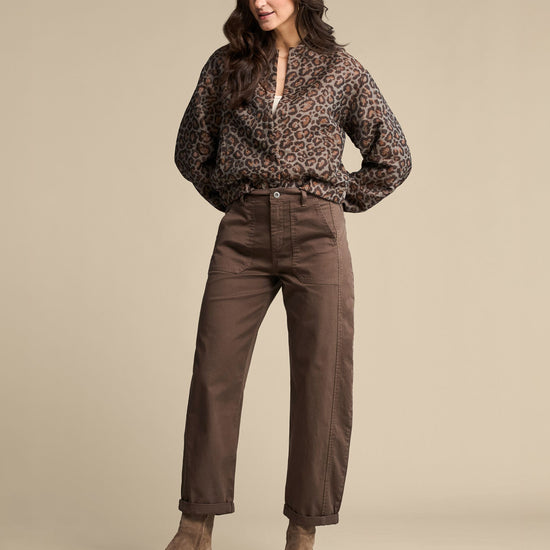 A woman modeling a brown leopard-print long-sleeve blouse and brown straight-leg pants, paired with taupe ankle boots, against a solid beige studio background.