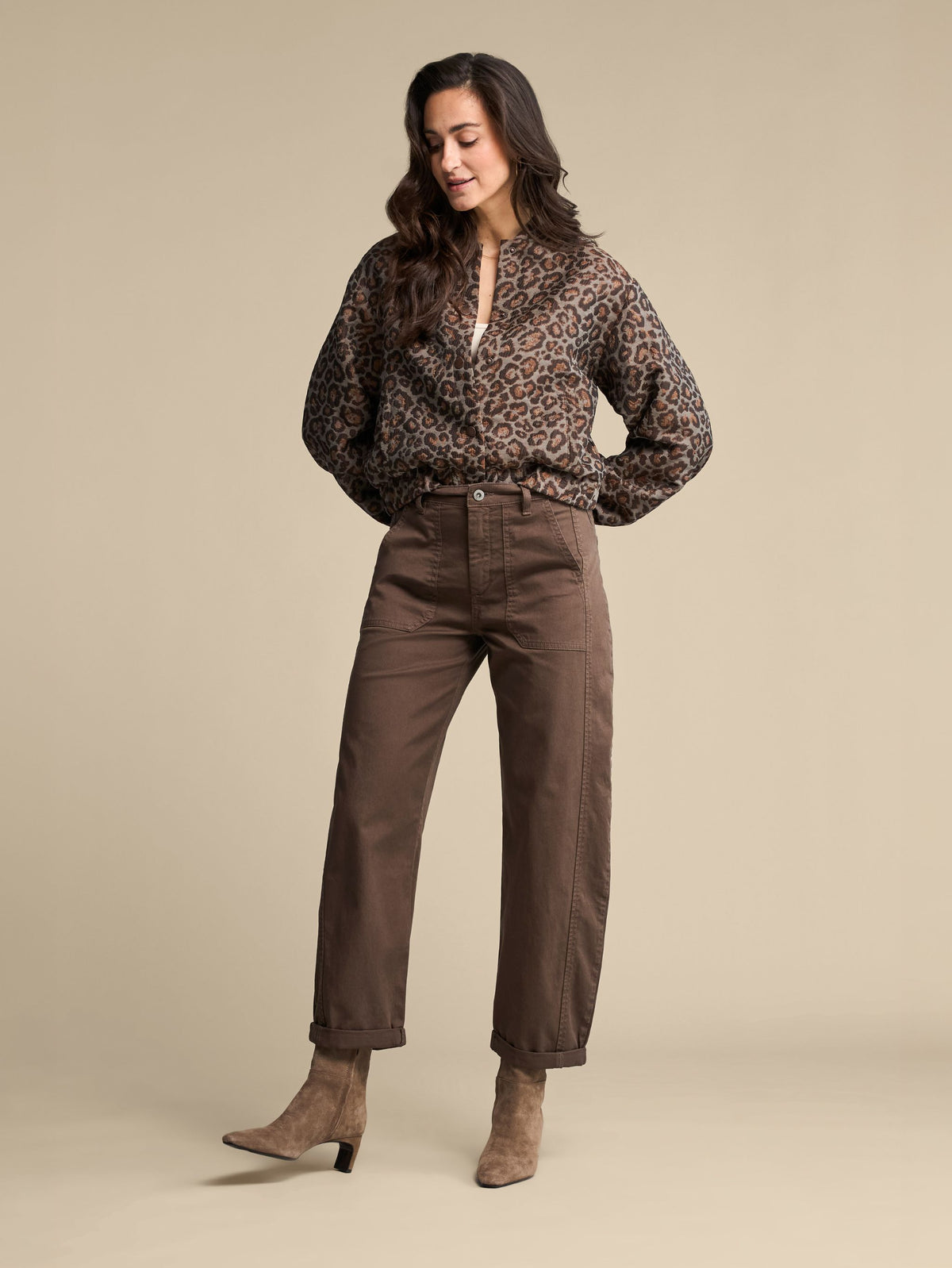 A woman modeling a brown leopard-print long-sleeve blouse and brown straight-leg pants, paired with taupe ankle boots, against a solid beige studio background.