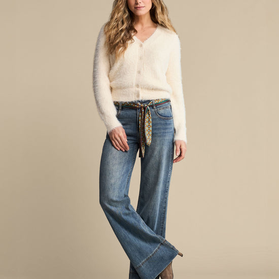 A woman modeling a cream fuzzy knit cardigan and blue wide-leg jeans with a patterned tie belt, standing against a solid beige studio background.