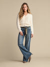 A woman modeling a cream fuzzy knit cardigan and blue wide-leg jeans with a patterned tie belt, standing against a solid beige studio background.