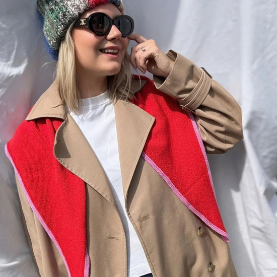 A woman wearing a beige trench coat with a bold red triangular scarf with pink trim posing against a white draped backdrop. 