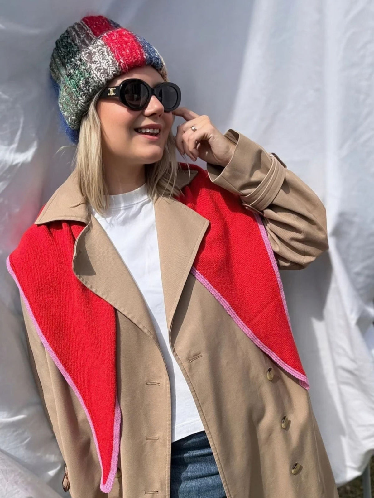 A woman wearing a beige trench coat with a bold red triangular scarf with pink trim posing against a white draped backdrop. 