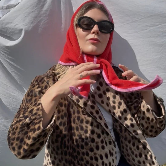 A woman wearing a red and pink patterned triangular scarf, black sunglasses, and a leopard print blazer, posing in front of a white draped backdrop.