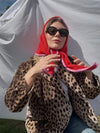 A woman wearing a red and pink patterned triangular scarf, black sunglasses, and a leopard print blazer, posing in front of a white draped backdrop.
