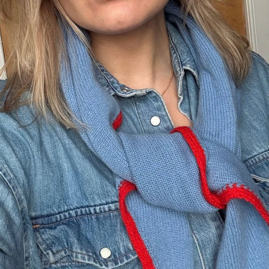 A close-up of a person wearing a light blue knit scarf with bold red trim, styled over a denim shirt.