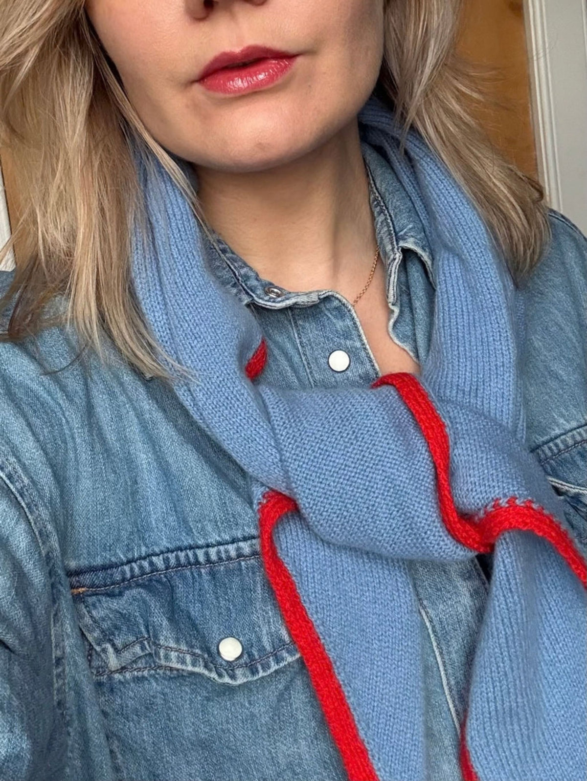 A close-up of a person wearing a light blue knit scarf with bold red trim, styled over a denim shirt.