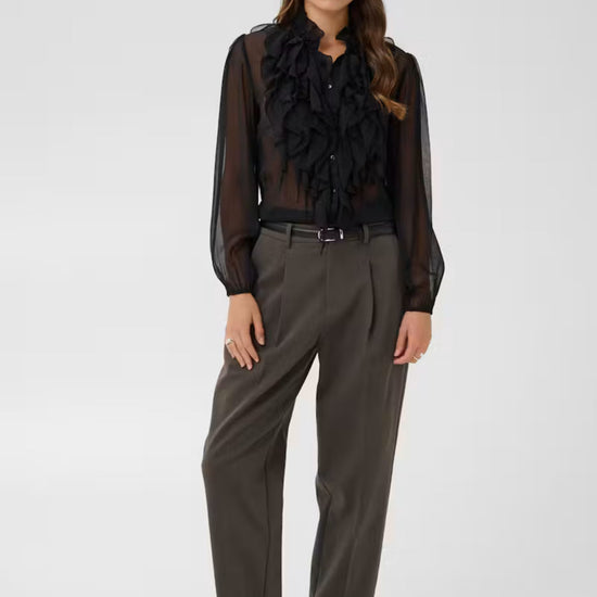 A woman modeling a sheer black long-sleeve blouse with dramatic ruffle detailing at the collar, paired with dark gray wide-leg trousers and black flats, against a light gray studio background.