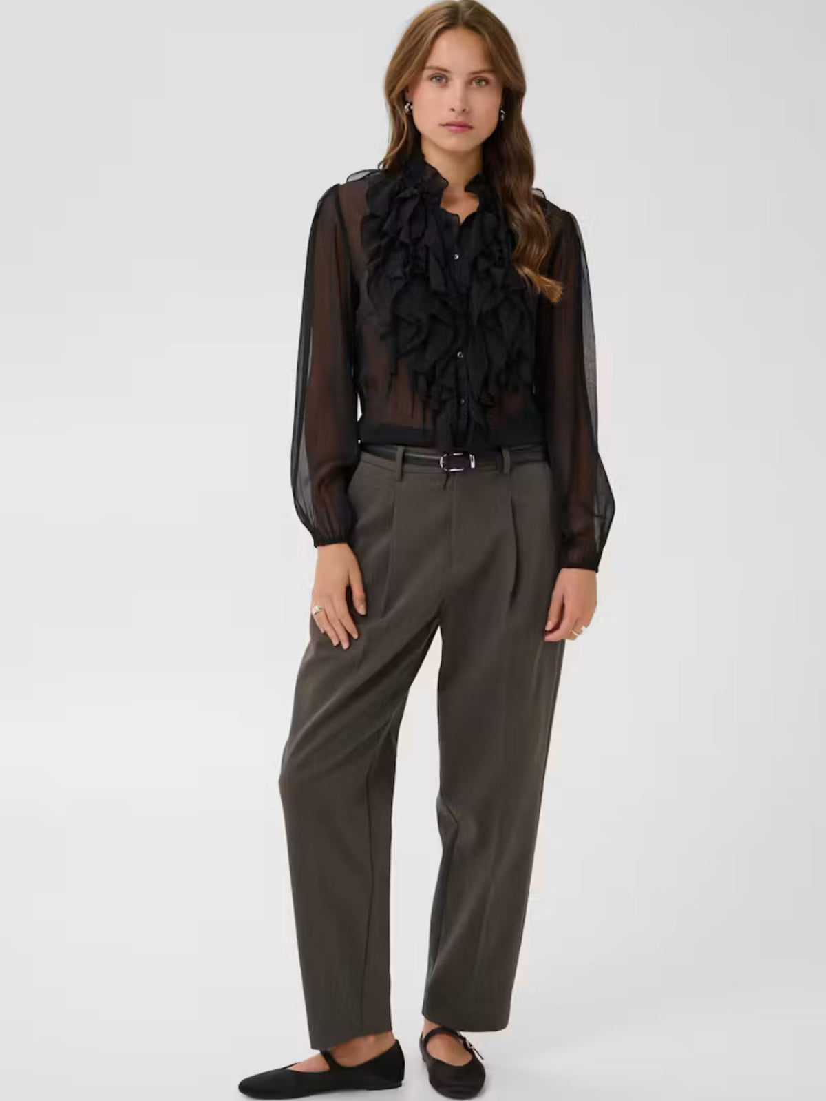A woman modeling a sheer black long-sleeve blouse with dramatic ruffle detailing at the collar, paired with dark gray wide-leg trousers and black flats, against a light gray studio background.