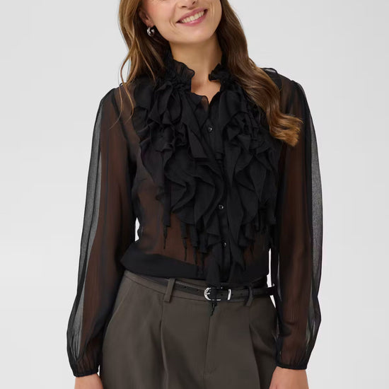 A woman wearing a sheer black long-sleeve blouse with a large ruffled collar and button front, paired with dark gray trousers, against a light gray studio background.