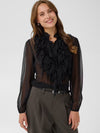 A woman wearing a sheer black long-sleeve blouse with a large ruffled collar and button front, paired with dark gray trousers, against a light gray studio background.