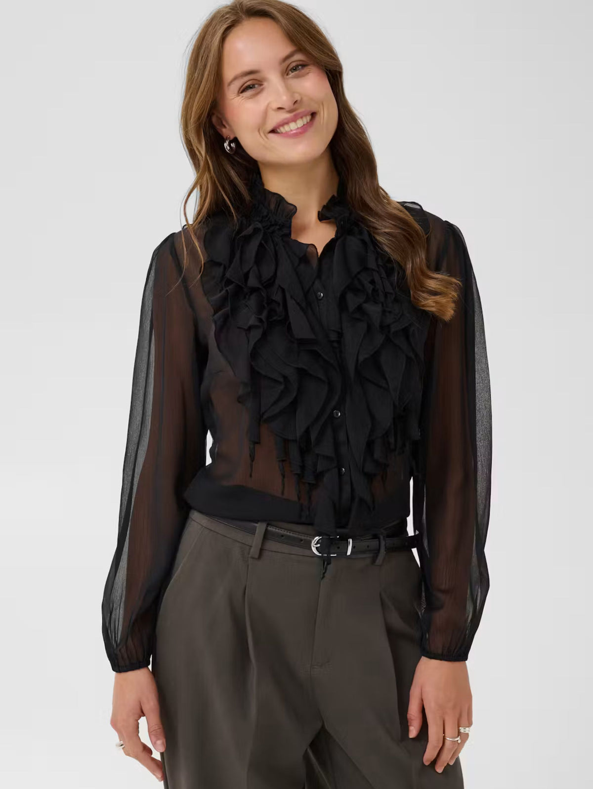 A woman wearing a sheer black long-sleeve blouse with a large ruffled collar and button front, paired with dark gray trousers, against a light gray studio background.