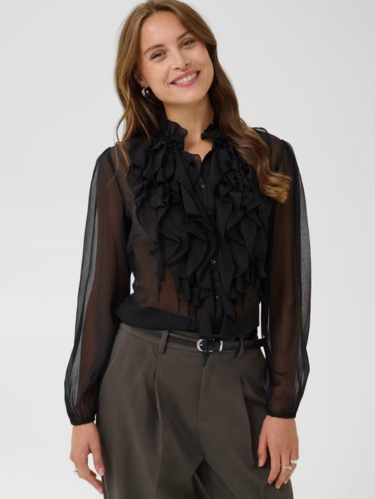A woman wearing a sheer black long-sleeve blouse with a large ruffled collar and button front, paired with dark gray trousers, against a light gray studio background.