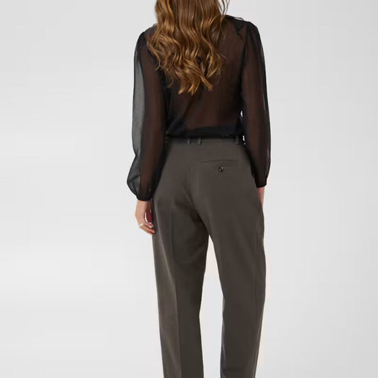 A woman shown from the back wearing a sheer black long-sleeve blouse and dark olive gray trousers, standing against a light gray studio background.