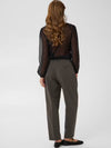 A woman shown from the back wearing a sheer black long-sleeve blouse and dark olive gray trousers, standing against a light gray studio background.