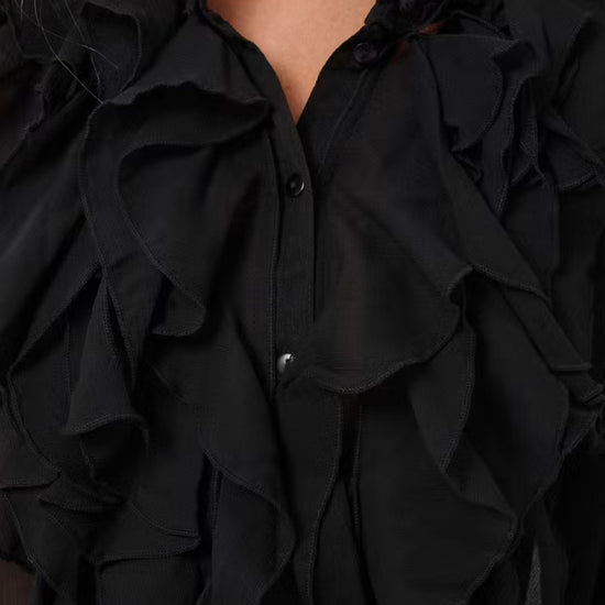 Close-up of a black ruffled blouse with layered cascading ruffles, a split neckline, and dark buttons, shown on a model.