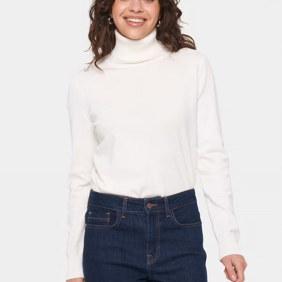 A woman wearing a white long-sleeve turtleneck sweater and dark blue jeans, standing against a light gray studio background.