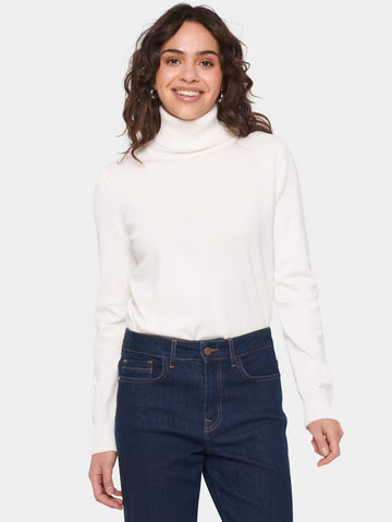 A woman wearing a white long-sleeve turtleneck sweater and dark blue jeans, standing against a light gray studio background.