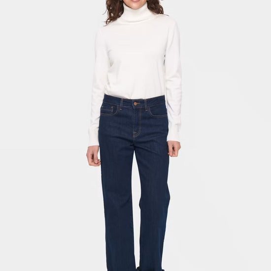 A woman modeling a white long-sleeve turtleneck sweater and dark blue straight-leg jeans with rolled cuffs, paired with brown suede boots, against a light gray background.