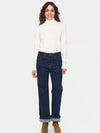 A woman modeling a white long-sleeve turtleneck sweater and dark blue straight-leg jeans with rolled cuffs, paired with brown suede boots, against a light gray background.