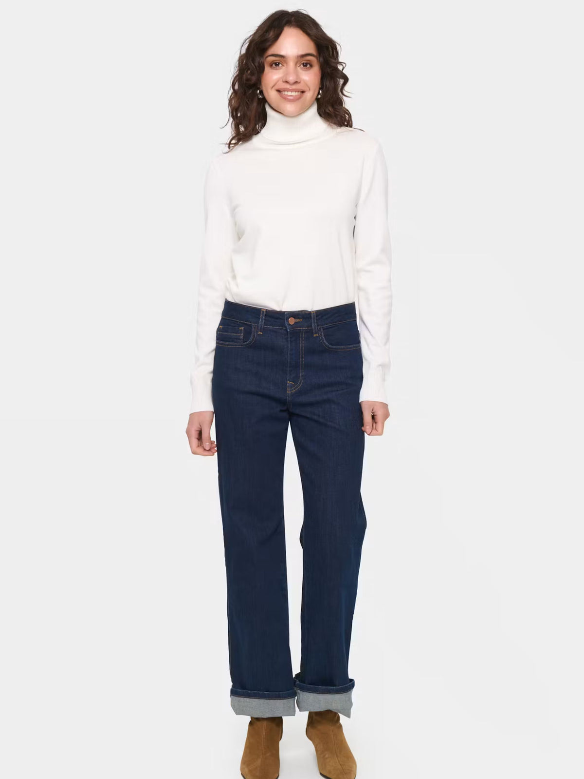 A woman modeling a white long-sleeve turtleneck sweater and dark blue straight-leg jeans with rolled cuffs, paired with brown suede boots, against a light gray background.