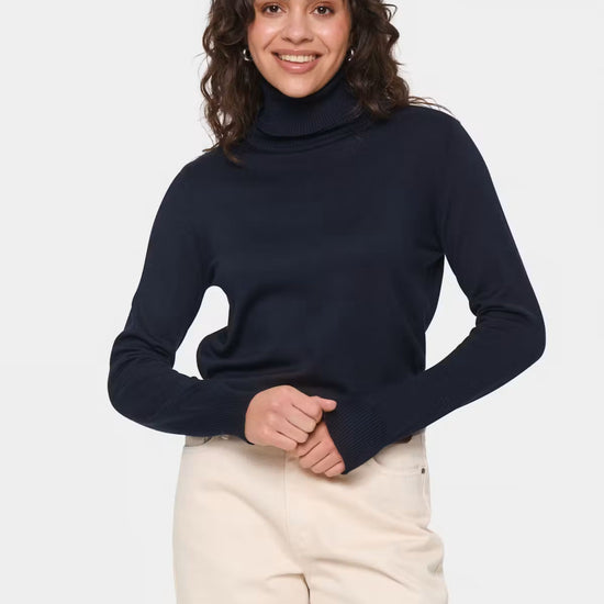 A woman wearing a dark navy blue ribbed turtleneck sweater and cream-colored trousers, standing against a light gray studio background.