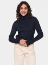A woman wearing a dark navy blue ribbed turtleneck sweater and cream-colored trousers, standing against a light gray studio background.
