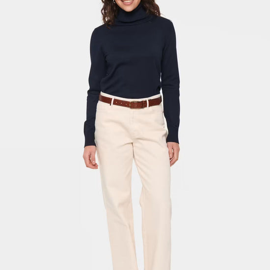 A woman modeling a dark navy turtleneck sweater and straight-leg cream trousers with a brown belt, paired with leopard print flats, against a light gray studio background.