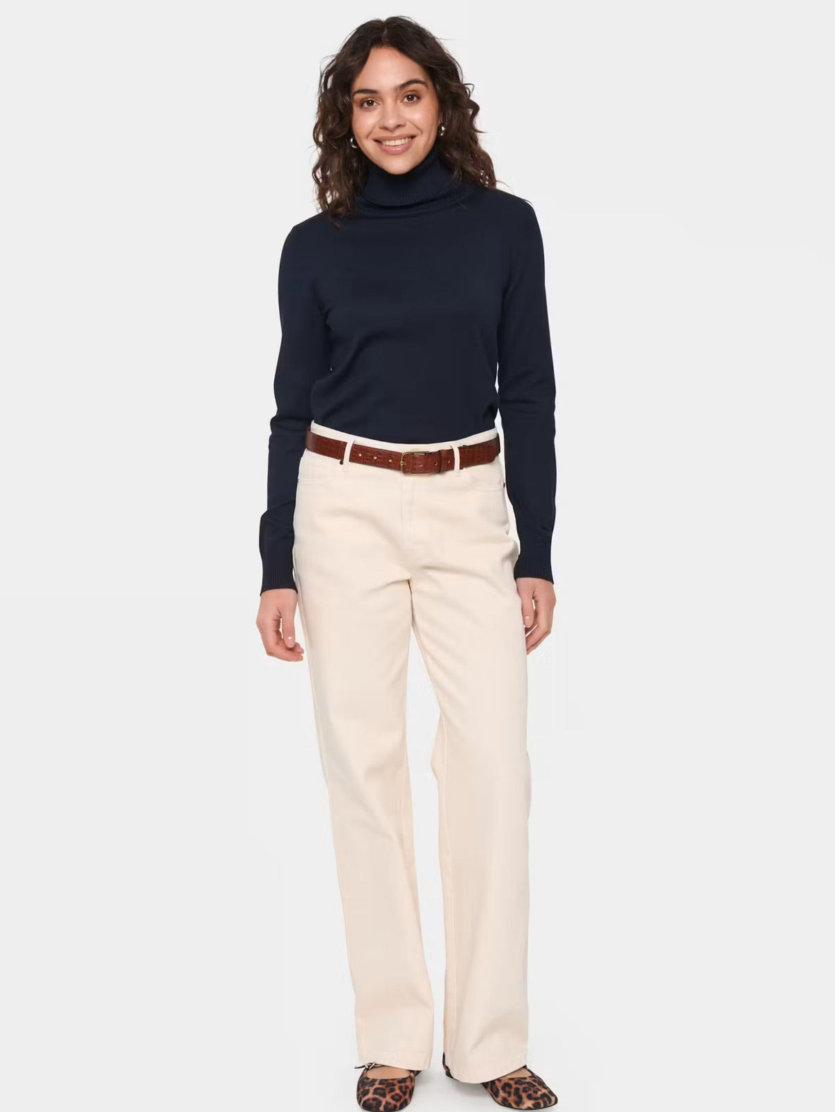 A woman modeling a dark navy turtleneck sweater and straight-leg cream trousers with a brown belt, paired with leopard print flats, against a light gray studio background.