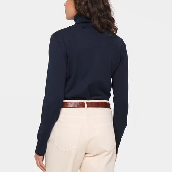 A woman shown from the back wearing a dark navy long-sleeve turtleneck sweater, cream-colored trousers, and a brown leather belt, against a light gray studio background.