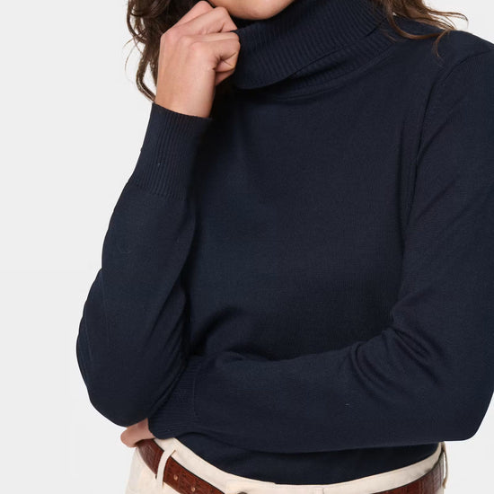 A woman wearing a dark navy ribbed turtleneck sweater, cream pants, and a brown belt, shown from the chest up against a light gray studio background.