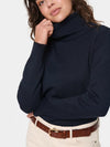 A woman wearing a dark navy ribbed turtleneck sweater, cream pants, and a brown belt, shown from the chest up against a light gray studio background.