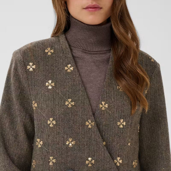 A close-up of a woman wearing a textured brown double-breasted blazer with small gold embroidered floral motifs, layered over a dark brown ribbed turtleneck sweater.