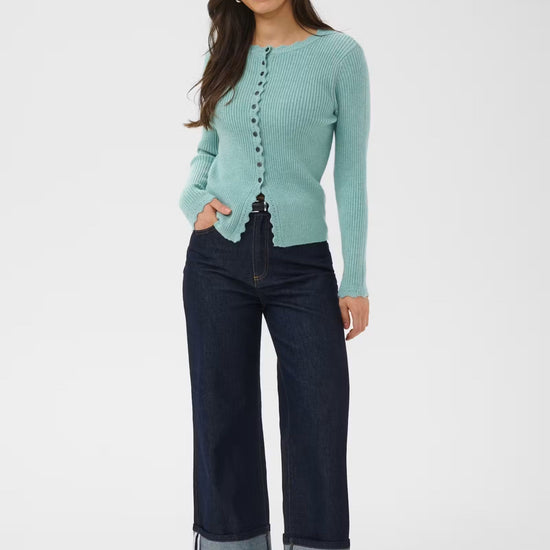A woman modeling a light aqua ribbed knit cardigan with front buttons, paired with dark blue wide-leg jeans with rolled cuffs and black loafers, against a plain white studio background.