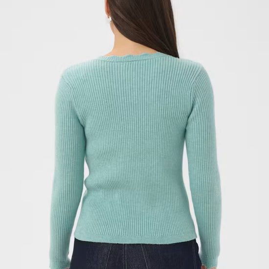 A woman viewed from the back wearing a ribbed long-sleeve sweater in a light aqua color, paired with dark blue jeans, against a plain light gray studio background.