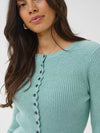 A close-up of a woman wearing a aqua ribbed knit cardigan sweater with dark buttons down the front, set against a plain light gray background.