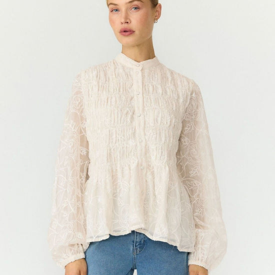 A woman modeling a cream-colored, long-sleeved lace blouse with a high mock neck and gathered peplum hem, paired with blue jeans, against a plain white studio background.