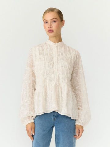 A woman modeling a cream-colored, long-sleeved lace blouse with a high mock neck and gathered peplum hem, paired with blue jeans, against a plain white studio background.