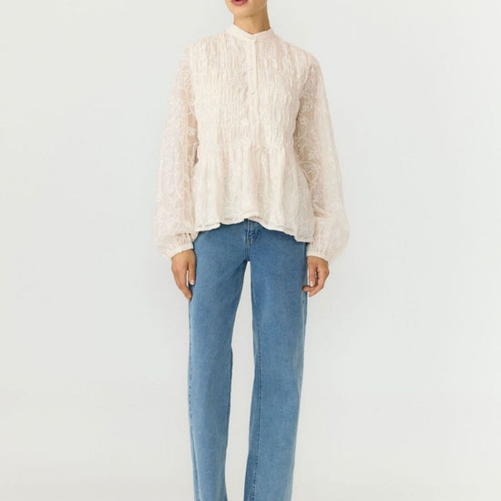A model stands against a plain light gray studio background wearing a cream-colored, textured long-sleeve blouse with a high mock neck, paired with light-wash blue jeans and black pointed-toe heels.