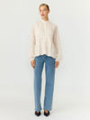 A model stands against a plain light gray studio background wearing a cream-colored, textured long-sleeve blouse with a high mock neck, paired with light-wash blue jeans and black pointed-toe heels.