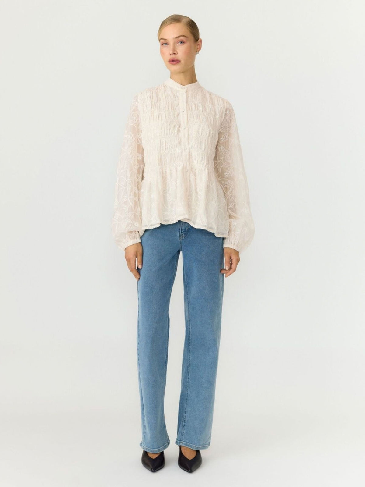 A model stands against a plain light gray studio background wearing a cream-colored, textured long-sleeve blouse with a high mock neck, paired with light-wash blue jeans and black pointed-toe heels.