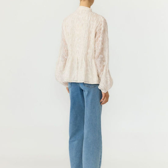 A woman viewed from the back wearing a cream-colored textured lace blouse with balloon sleeves and light blue wide-leg jeans, set against a plain white studio background.