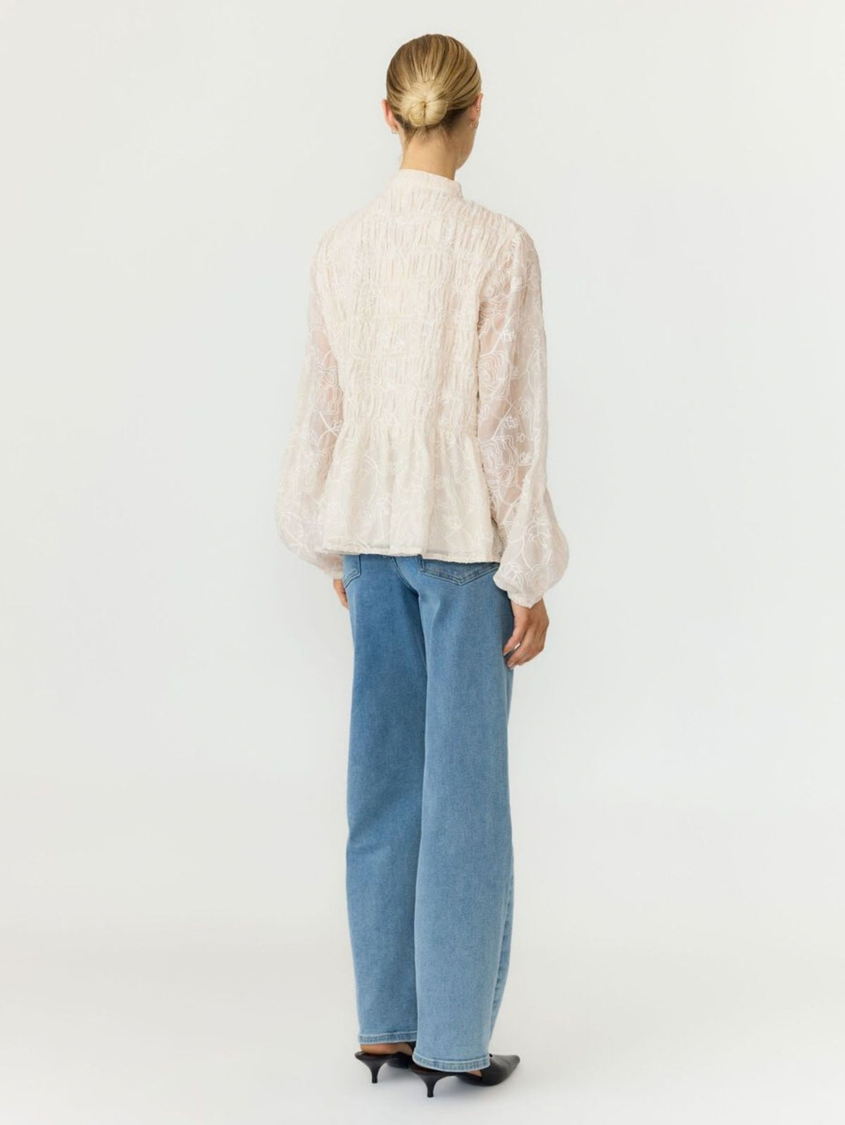 A woman viewed from the back wearing a cream-colored textured lace blouse with balloon sleeves and light blue wide-leg jeans, set against a plain white studio background.